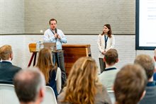 Roderick Kleij praat door een microfoon terwijl Isabel Leemhuis toekijkt. Ze staan voor de zaal.